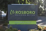 A gray sign stands in front of some trees and a roadway. The sign has green text that says Rosboro.