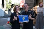Oregon Governor Kate Brown poses with a man to her right in Japan. They hold a "state of Oregon" sign together and give thumbs ups.