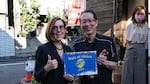 Oregon Governor Kate Brown poses with a man to her right in Japan. They hold a "state of Oregon" sign together and give thumbs ups.