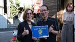 Oregon Governor Kate Brown poses with a man to her right in Japan. They hold a "state of Oregon" sign together and give thumbs ups.