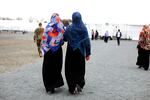 Two women wearing headscarves walk with their backs to the camera across a gravel road.