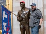 A ma stands on the base of a statue in the US Capitol building. He has put a red "Make American Great Again" on the statute, and a "Trump" flag in the statue's hands.