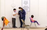 Children and adults stand or sit in chairs in a room. A sign on the wall behind them says "COVID-19 accine recovery area."