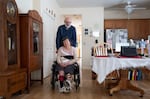 A man with a gray beard has his arms on the back of the wheelchair of a woman with short hair. They are in a dining room with elegant wood furniture and a table covered in a white table cloth.