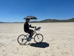 A man wearing a floppy hat and holding an umbrella rides a bicycle over a parched desert landscape.