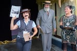Liz Bartholomew (left) wins for best Buster Keaton look-alike after being awarded by Dana Merryday (center) and Jennifer Ferraez Chartrand during a 1920s themed costume contest. Liz is wearing a porkpie hat, a fake mustache, a vest over a striped short-sleeve button down shirt and carrying magnifying glass and a handwritten sign that says "How to be a detective." Dana is wearing a gray vintage suit and straw hat while Jennifer is wearing a green 1920s-style beaded dress and has a headband with a peacock feather in it.