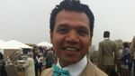 A man wears a tan suit with a flower pinned to his lapel and smiles at the camera, at what appears to be a formal attire outdoor event. Behind him, people dressed in nice clothes walk on a grassy lawn between tents and awnings.