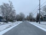 To the right, two people walk down the sidewalk of a residential street on a cold winter day. The sky is cloudy and the sides of the road have a dusting of snow.