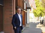 Dressed in a business casual suit, Drotzmann stands in front of brick and wood paneled store fronts on a sunny day