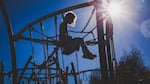 Little girl with curly hair sits on top of a playground jungle gym.