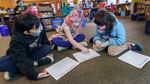 Students sit together on the floor of a classroom wearing face masks and looking at worksheets.