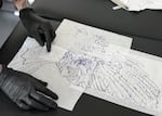 On a black table, hands wearing black gloves are seen arranging three pieces of paper to make up a drawing of an eagle in black ink.