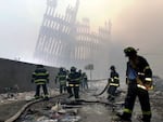 Firefighters wearing helmets and dark uniforms with shining reflective strips work at the site of the destroyed World Trade Center in New York City on September 11, 2001. Debris litters the ground, and the partial skeleton of one building rises in the hazy background.