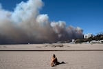 A lone sunbather sits on the beach and watches a large plume of smoke from a wildfire rise over the Pacific Palisades.