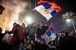 Crowds of people celebrate in Belgrade’s Slavija Square on October 25, 2026, after Aleksandar Vučić conceded defeat in Serbia's parliamentary elections. The people are waving flags and holding up their hands. Fireworks explode in the darkened sky.