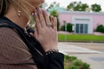 A woman clasps in hands in prayer, behind her is the pink house.