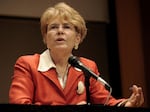 A woman in an orange suit jacket and white blouse gestures as she sits at a table with a microphone.