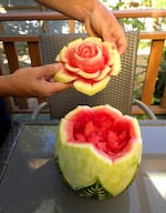 A pair of hands hold up a chunk of watermelon carved to look like a flower. The rest of the watermelon is cut open to reveal the fruit inside, but further down the rind are carvings etched to look like leaves.