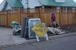 A dirt-streaked warning sign leans against a washing machine and other assorted debris on the side of the road.