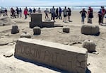 Several sand sculptures are behind a sand block sign reading "PUGKIN PATCH." There are pumpkins with the faces of pug dogs, and a wagon with hay bales on it. Spectators walk by on the beach in the background.