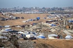 In this photo, a tent camp stands amid destroyed buildings. The tents are made primarily from white and blue tarps. Piles of rubble rise up between some clusters of tents.