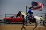 A cowboy rides a horse inside a fenced outdoor arena. An American flag is displayed largely on the wall behind him.