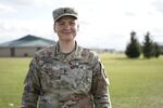 A person in camouflage military clothing smiles at the camera with a grassy lawn in the background.