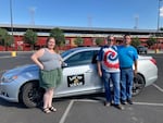 Two women and a man stand in front of a silver sedan in a parking lot. The car has a poster on the side with the Let'er Uber logo on it.