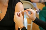 A woman in a black shirt gets a shot in her upper arm. All you can see are the patient's shoulder and the provider's hands.