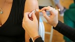 A woman in a black shirt gets a shot in her upper arm. All you can see are the patient's shoulder and the provider's hands.