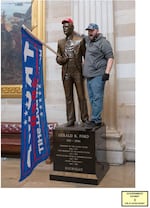 A ma stands on the base of a statue in the US Capitol building. He has put a red "Make American Great Again" on the statute, and a "Trump" flag in the statue's hands.