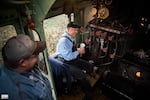 In this provided photo from Holiday Express  in December 2017, a volunteer looks on as Pat Tracy sits in the cab of the Southern Pacific 4449 steam locomotive. Pat Tracy wears a blue shirt and black overalls, the cab is mostly black with red dials dotted everywhere.