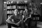 A black and white photo of co-authors Ben Hodgson and Laura Moulton. They're seated in a room together, with Hodgson on the left grinning at the camera, and Moulton on the right smiling at Hodgson. They're both wearing matching Street Books t-shirts with a silhouette of the bike library on them. Behind them is a large bookself full of books, standing lamps on either side, and photos on the wall.