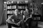 A black and white photo of co-authors Ben Hodgson and Laura Moulton. They're seated in a room together, with Hodgson on the left grinning at the camera, and Moulton on the right smiling at Hodgson. They're both wearing matching Street Books t-shirts with a silhouette of the bike library on them. Behind them is a large bookself full of books, standing lamps on either side, and photos on the wall.