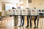 Voters in Detroit stand in voting booths that say "Vote" with an American flag on the side. We can see their legs and feet but not their faces, which are obscured by the voting booth. 