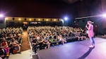 Visible in the foreground is a women, Ayleen Crotty,  in a red dress and blue tights speaking to a theatre audience of about 300 people.