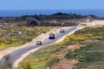 In this photo, three military vehicles drive along a road. The sides of the paved road have green foliage, yellow flowers, patches of brown dirt and piles of debris.