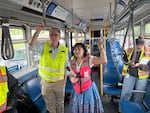A white man in a yellow vest stands in the bus next to a shorter dark-haired woman with a pink vest and dress.