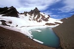 snow melts off a craggy mountain peak into a blue-green lake encircled by a hiking trail