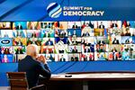 Joe Biden sits at a table with his back to the camera, facing a wall-sized screen with images of multiple people in a rectangular grid. The words "Summit for Democracy" appear at the top of the screen.