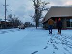 Two people walk down a snow-covered sidewalk, leaving behind footprints.