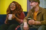 Deltra James and Mike Carbone sit next to each other on a couch holding coffee cups. Deltra is laughing and Mike is looking at her.