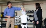 A man in a blue stands with his hand on the head of a white humanoid robot. A woman in a black blouse stands on the other side of the robot.