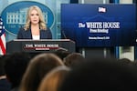 In this photo, White House press secretary Karoline Leavitt is standing behind a lectern with two microphones attached to it. She is wearing a dark blue blazer with light pinstripes on it. On the right is a large TV screen that says "The White House Press Briefing" on it.