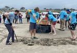 Seven people work on various parts of the beginning of a sand sculpture. One pours ocean water into a sand form the center, while several more rake sand in the foreground. Most of them are wearing blue event t-shirts.
