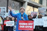 A crowd holding signs, including one that reads "Hands off our CFPB," joins Sen. Elizabeth Warren in front of the Consumer Financial Protection Bureau.