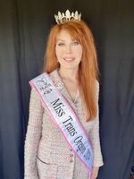 A woman with long red hair in a pink suit poses in a sash and crown.