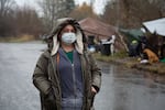 Jamie Spinelli stands bundled up in warm clothes and a mask, in front of a few tents at a homeless camp in Clark County. The weather is rainy.