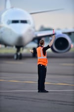 A man in an orange vest gestures to an airplane in the background.