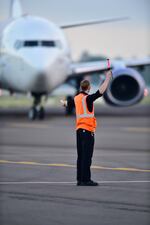 A man in an orange vest gestures to an airplane in the background.